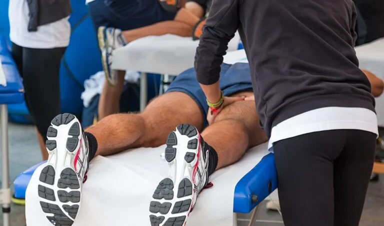 A person is lying face down on a massage table receiving a leg massage, with a massage therapist working on their thigh. Several other massage tables and people are visible in the background.